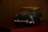 A vintage toy car model, mainly in green and silver with a classic design, placed on a dark surface against a blurred background. It appears to be a miniature of a classic sedan, capturing the details of the headlights and front grille.