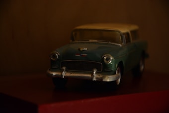 A vintage toy car model, mainly in green and silver with a classic design, placed on a dark surface against a blurred background. It appears to be a miniature of a classic sedan, capturing the details of the headlights and front grille.