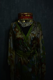 A mannequin wearing a colorful, patterned robe with a wide, ornate red choker adorned with gold coins and pearls. The fabric of the robe features swirling designs in shades of green, brown, and beige. The background is dark, enhancing the contrast of the vibrant clothing.