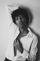 A black and white portrait of a person with short, curly hair wearing a white open shirt. They are posing with one hand raised behind their head, striking a confident and expressive pose against a plain background.