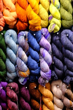 A colorful assortment of yarn skeins is arranged in a grid pattern. The skeins are twisted and feature a wide range of colors including shades of orange, yellow, green, blue, purple, and brown. The texture of the yarn is clearly visible, giving a sense of softness and warmth.