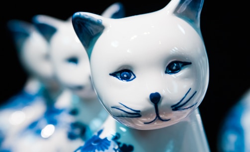 A close-up of a porcelain cat figurine with intricate blue designs on its ears, eyes, and body. The cat has a serene expression and glossy finish, with other similar figurines blurred in the background.