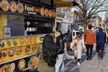 A bustling street scene featuring a food truck offering halal options. The yellow menu on the truck displays various dishes. Several people walk past, dressed in winter attire, while one person stands in line, approaching the truck's counter. Buildings and storefronts line the street in the background, with a tree visible on the right side.