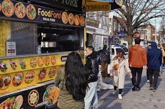 A bustling street scene featuring a food truck offering halal options. The yellow menu on the truck displays various dishes. Several people walk past, dressed in winter attire, while one person stands in line, approaching the truck's counter. Buildings and storefronts line the street in the background, with a tree visible on the right side.