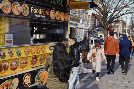 A bustling street scene featuring a food truck offering halal options. The yellow menu on the truck displays various dishes. Several people walk past, dressed in winter attire, while one person stands in line, approaching the truck's counter. Buildings and storefronts line the street in the background, with a tree visible on the right side.
