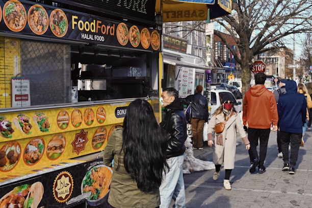A bustling street scene featuring a food truck offering halal options. The yellow menu on the truck displays various dishes. Several people walk past, dressed in winter attire, while one person stands in line, approaching the truck's counter. Buildings and storefronts line the street in the background, with a tree visible on the right side.