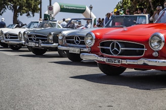 Multiple classic Mercedes-Benz cars are lined up on a street with people gathered around, possibly for a car show or event. The cars are in a range of colors, including red, white, and silver, and the scene is set in a sunny outdoor environment.