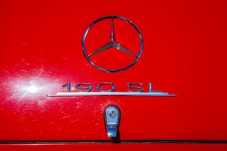 A close-up of a classic car's rear, prominently featuring a chrome Mercedes-Benz logo above the model designation '190 SL' and a chrome handle against a vibrant red background.