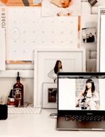 A workspace is depicted with a laptop displaying a magazine cover featuring a woman. Above the laptop, there is a calendar showing the month of October. There are framed photos, including an illustration of a woman and a baby photograph on the wall. The desk holds a keyboard and various bottles, including one with a pump dispenser.