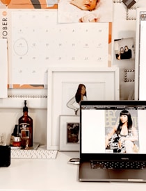 A workspace is depicted with a laptop displaying a magazine cover featuring a woman. Above the laptop, there is a calendar showing the month of October. There are framed photos, including an illustration of a woman and a baby photograph on the wall. The desk holds a keyboard and various bottles, including one with a pump dispenser.