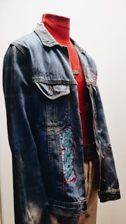 A denim jacket displayed on a mannequin, featuring embroidered patches and paired with a high-necked red shirt underneath. The setup is well-lit, highlighting the textures and colors of the jacket and shirt.
