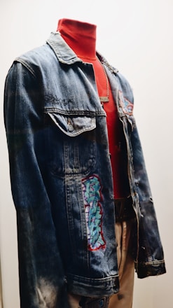 A denim jacket displayed on a mannequin, featuring embroidered patches and paired with a high-necked red shirt underneath. The setup is well-lit, highlighting the textures and colors of the jacket and shirt.