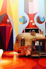 An elaborate, artistic installation featuring a whimsical, metallic caravan with a human face on it. The walls around the artwork display vibrant geometric patterns and colors. A mannequin dressed in colorful clothing stands on a red triangular platform. Lamps and various objects adorn the top of the caravan, contributing to the surreal and eclectic aesthetic.