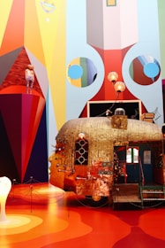 An elaborate, artistic installation featuring a whimsical, metallic caravan with a human face on it. The walls around the artwork display vibrant geometric patterns and colors. A mannequin dressed in colorful clothing stands on a red triangular platform. Lamps and various objects adorn the top of the caravan, contributing to the surreal and eclectic aesthetic.