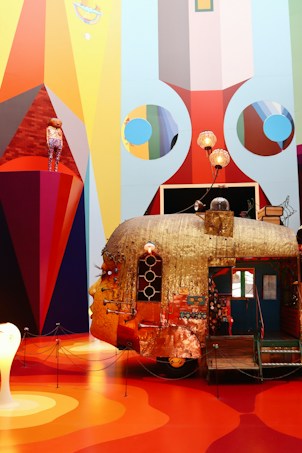 An elaborate, artistic installation featuring a whimsical, metallic caravan with a human face on it. The walls around the artwork display vibrant geometric patterns and colors. A mannequin dressed in colorful clothing stands on a red triangular platform. Lamps and various objects adorn the top of the caravan, contributing to the surreal and eclectic aesthetic.
