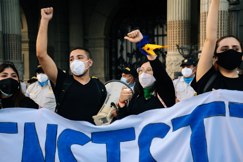 homem no colete preto vestindo máscara branca segurando bandeira branca e azul