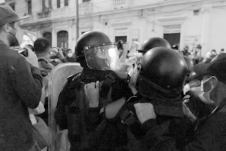 A black and white image captures a group of people standing close together, some wearing helmets and face masks. Several individuals appear to be holding shields or using cameras to record. The scene seems to take place in an urban environment with a building facade in the background.