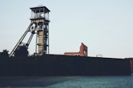 An industrial scene featuring a tall, metal mining tower or headframe. In the background, there are aged brick buildings and minimal vegetation. The architecture suggests industrial heritage with a focus on mining.