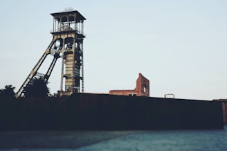 An industrial scene featuring a tall, metal mining tower or headframe. In the background, there are aged brick buildings and minimal vegetation. The architecture suggests industrial heritage with a focus on mining.