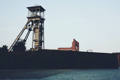 An industrial scene featuring a tall, metal mining tower or headframe. In the background, there are aged brick buildings and minimal vegetation. The architecture suggests industrial heritage with a focus on mining.