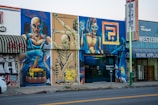 A colorful street mural featuring three futuristic, muscular figures in dynamic poses, painted on a city building facade. The largest figure holds a drumstick, another is suspended in mid-air, and the third wears goggles, all set against a vivid urban fantasy background. Graffiti art is interspersed with commercial shop signs.