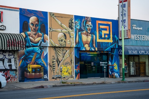 A colorful street mural featuring three futuristic, muscular figures in dynamic poses, painted on a city building facade. The largest figure holds a drumstick, another is suspended in mid-air, and the third wears goggles, all set against a vivid urban fantasy background. Graffiti art is interspersed with commercial shop signs.