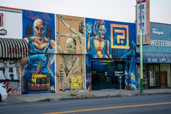 A colorful street mural featuring three futuristic, muscular figures in dynamic poses, painted on a city building facade. The largest figure holds a drumstick, another is suspended in mid-air, and the third wears goggles, all set against a vivid urban fantasy background. Graffiti art is interspersed with commercial shop signs.
