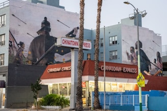 A restaurant named Chinatown Express is situated on a street corner, with a large mural on the adjacent building depicting historical figures in dramatic poses, possibly from a battle scene. The mural includes people in traditional clothing with arrows flying through the air. Palm trees are visible in the foreground, and the sky is overcast.