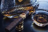 A close-up of a dark brown cigar resting on a metal stand, placed next to a glass of amber liquid likely whisky. In the background, a crystal decanter filled with a similar amber liquid is visible, featuring intricate patterns.