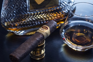 A close-up of a dark brown cigar resting on a metal stand, placed next to a glass of amber liquid likely whisky. In the background, a crystal decanter filled with a similar amber liquid is visible, featuring intricate patterns.