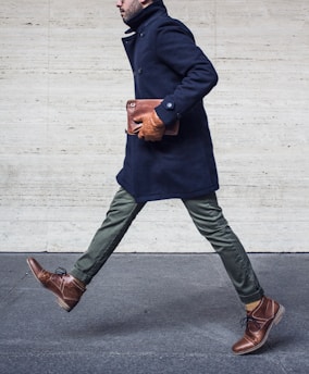 A person in a stylish navy coat and green pants is walking with a determined stride. They are wearing brown leather shoes and gloves, holding a brown leather clutch under their arm against a neutral stone wall background.