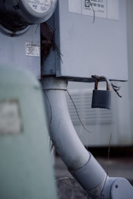 An outdoor electrical meter box attached to a wall, with a thick gray conduit pipe leading downwards. A rusty padlock is affixed to a latch, providing a sense of security. There are warning labels and some exposed wires visible.