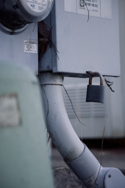 An outdoor electrical meter box attached to a wall, with a thick gray conduit pipe leading downwards. A rusty padlock is affixed to a latch, providing a sense of security. There are warning labels and some exposed wires visible.