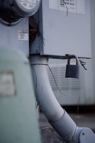 An outdoor electrical meter box attached to a wall, with a thick gray conduit pipe leading downwards. A rusty padlock is affixed to a latch, providing a sense of security. There are warning labels and some exposed wires visible.
