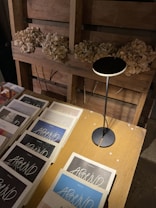 A table is set against a wooden backdrop adorned with dried flowers. On the table, there is a collection of magazines or books titled 'AROUND', featuring various cover designs. A sleek, modern lamp casts light over the scene.
