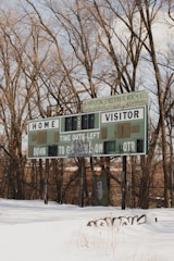 An old scoreboard stands in a snowy field, surrounded by leafless trees. The structure appears weathered, with patches of graffiti on it. The sky is partly cloudy, and the snow on the ground is undisturbed, except for some grass stalks poking through.