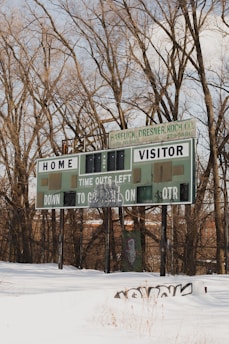 An old scoreboard stands in a snowy field, surrounded by leafless trees. The structure appears weathered, with patches of graffiti on it. The sky is partly cloudy, and the snow on the ground is undisturbed, except for some grass stalks poking through.