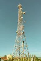A tall communication tower equipped with various satellite dishes and antennas reaches into a clear blue sky. The tower is surrounded by a fence and appears to be located in a semi-rural area with green vegetation and small structures with red-tiled roofs nearby.