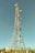 A tall communication tower equipped with various satellite dishes and antennas reaches into a clear blue sky. The tower is surrounded by a fence and appears to be located in a semi-rural area with green vegetation and small structures with red-tiled roofs nearby.
