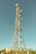 A tall communication tower equipped with various satellite dishes and antennas reaches into a clear blue sky. The tower is surrounded by a fence and appears to be located in a semi-rural area with green vegetation and small structures with red-tiled roofs nearby.