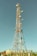 A tall communication tower equipped with various satellite dishes and antennas reaches into a clear blue sky. The tower is surrounded by a fence and appears to be located in a semi-rural area with green vegetation and small structures with red-tiled roofs nearby.