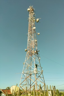 A tall communication tower equipped with various satellite dishes and antennas reaches into a clear blue sky. The tower is surrounded by a fence and appears to be located in a semi-rural area with green vegetation and small structures with red-tiled roofs nearby.
