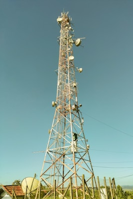A tall communication tower equipped with various satellite dishes and antennas reaches into a clear blue sky. The tower is surrounded by a fence and appears to be located in a semi-rural area with green vegetation and small structures with red-tiled roofs nearby.