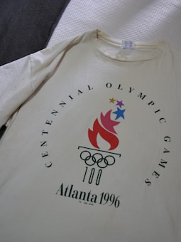 A cream-colored T-shirt displays the logo for the 1996 Atlanta Centennial Olympic Games featuring a stylized flame in red with stars above it and the Olympic rings below, surrounded by text.