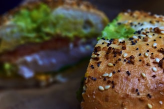 A close-up of a sesame seed bun sandwich with visible layers of ingredients, including fresh green lettuce. The focus is on the textured, golden-brown bun sprinkled with sesame and poppy seeds.