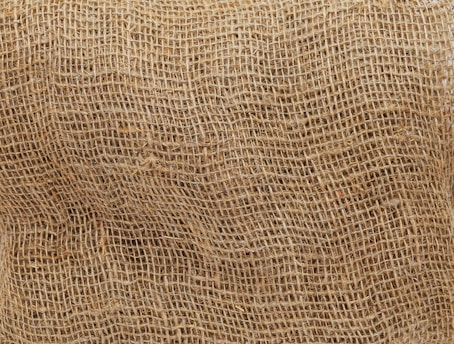 A close-up view of a brown burlap sack, highlighting its coarse and textured weave. The natural fibers create a rough surface pattern, characteristic of the rustic and utilitarian material.