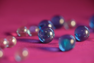 Various transparent and colored glass marbles are scattered on a vibrant pink surface, reflecting light and casting soft shadows. The marbles vary in size and display a mix of blue and clear colors, with some appearing slightly blurred in the background due to depth of field.