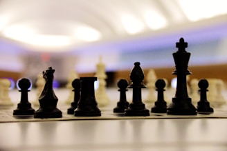 Chess pieces are arranged on a board in a strategic setup, viewed from a low angle. The focus is on the black pieces in the foreground with blurred white pieces in the background. The setting is softly lit with a modern and slightly futuristic atmosphere.