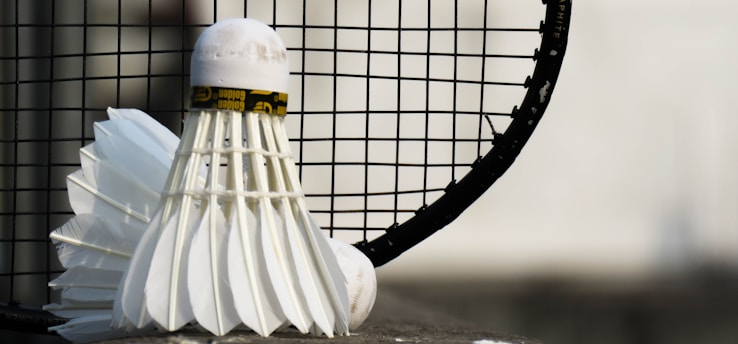 A close-up of a badminton racket striking a shuttlecock mid-air on an indoor court.