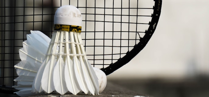 A close-up view of a shuttlecock leaning against a badminton racket with a focus on the net and feathers. The background is blurred, emphasizing the sports equipment in the foreground.